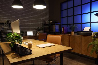 Karanlık ofisin içinde işyeri, bitkiler ve parlayan lambalar var.