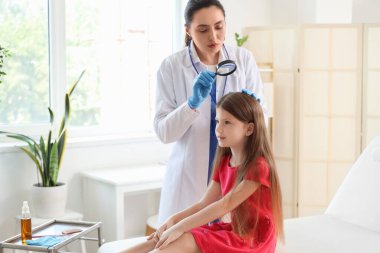Büyüteci olan kadın doktor klinikte küçük kızın kafasında pediküloz olup olmadığını kontrol ediyor.