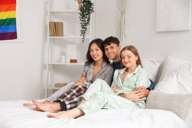 Young man with two women hugging in bedroom. Polyamory concept clipart