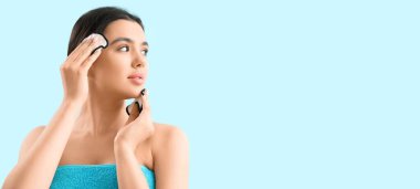 Açık mavi arkaplanda pamuk pedlerle makyaj malzemesi çıkaran genç bir kadın.