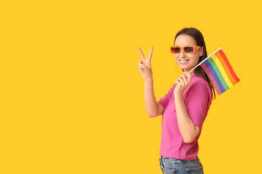 LGBT bayrağı taşıyan güneş gözlüklü güzel genç bir kadın sarı arka planda kalp hareketi gösteriyor.