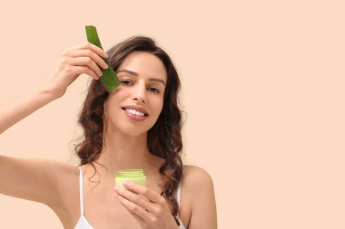 Krema kavanozu olan güzel genç bir kadın bej arka planda aloe vera yaprağı uyguluyor.