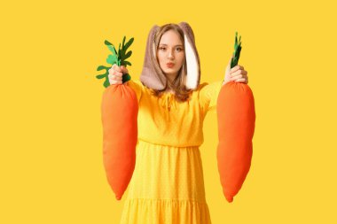 Sarı arka planda oyuncak havuçları olan tavşan kulaklı güzel bir kadın. Paskalya kutlaması