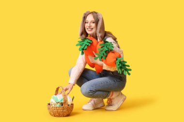 Tavşan kulaklı, oyuncak havuçlu ve sarı arka planda Paskalya sepetli güzel bir kadın.