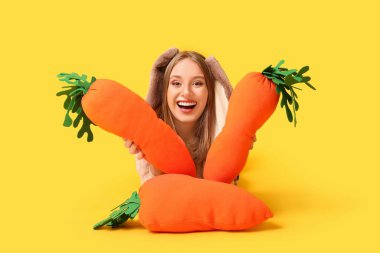 Sarı arka planda oyuncak havuçları olan tavşan kulaklı komik kadın. Paskalya kutlaması