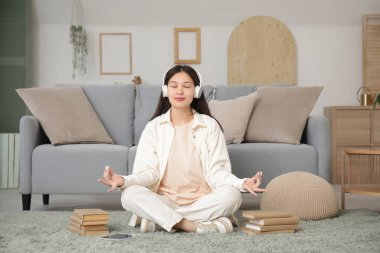 Kulaklıklı güzel bir Asyalı kadın oturma odasında halıda meditasyon yapıyor ve müzik dinliyor.