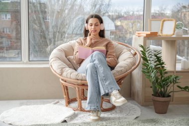 Kulaklıklı, kitapla sesli kitap dinleyen ve evdeki pencerenin yanındaki koltukta oturan güzel genç bir kadın.