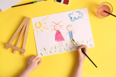 Çocuk eli, sarı arka planda fırça ve suluboya resimlerle insanları çiziyor.