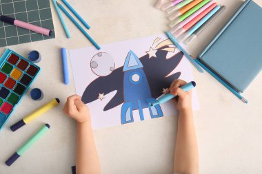 Beyaz grunge arkaplan üzerine işaretli roket, suluboya boyalar ve defterler çizen çocuk eli