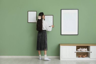 Genç bir kadın şık bir odada yeşil duvara resim çerçevesi asıyor.
