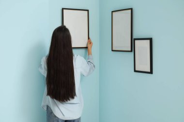 Genç bir kadın odadaki mavi duvara resim çerçevesi asıyor.