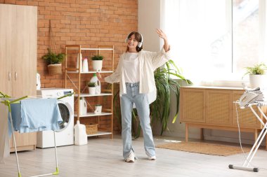 Kulaklıklı mutlu genç kadın çamaşırhanede dans ediyor.