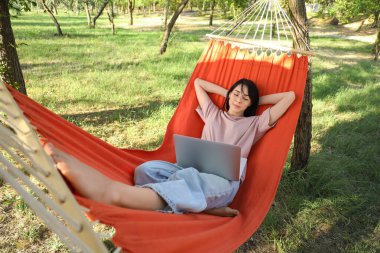 Bilgisayarlı genç bir kadın dışarıda hamakta dinleniyor.