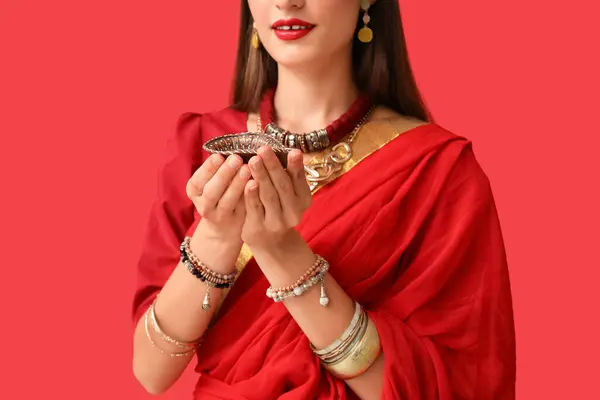 Beautiful young Indian woman in sari with diya lamp on red background, closeup