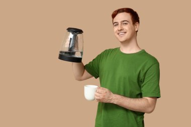 Modern elektrikli su ısıtıcısı ve bej arka planda fincanı olan yakışıklı genç adam.