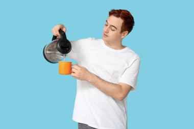 Yakışıklı genç adam modern elektrikli su ısıtıcısından mavi arka planda kupaya sıcak su döküyor.