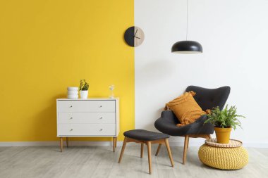 Şık oturma odasının içinde siyah koltuk, çekmeceler, lamba ve saat sarı duvarda.