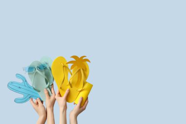 Mavi arka planda iki çift parmak arası terlik ve plaj aksesuarı tutan eller.