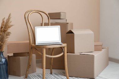 Sandalyede boş dizüstü bilgisayar. Odada bej duvarın yanında kutular var.