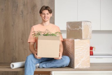 Elinde karton kutular ve bir rulo streç film olan genç adam mutfaktaki masada oturuyor.