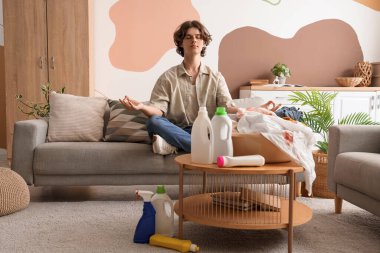 Kirli çamaşırları ve çamaşır deterjanıyla evde meditasyon yapan genç bir adam.