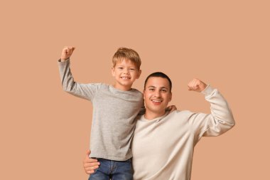 Mutlu baba ve küçük oğlu bej arka planda kaslarını gösteriyorlar.