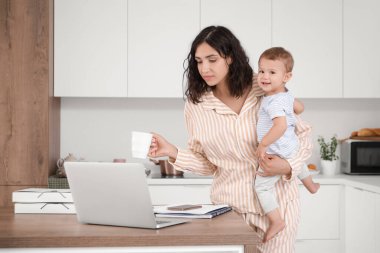 Kahve fincanı ve küçük bebeğiyle mutfakta çalışan bir anne.