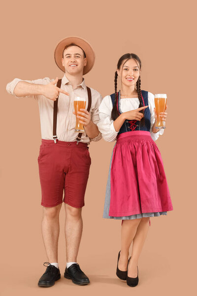 Молодая счастливая пара в традиционной немецкой одежде указывает на бокалы пива на бежевом фоне