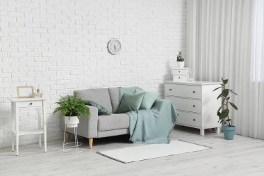 Gri kanepe, ev bitkileri, çekmeceler ve şık oturma odasındaki beyaz tuğlalı duvar üzerindeki saat.