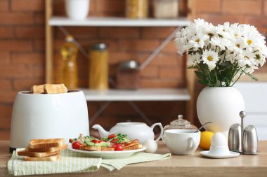 Çıtır ekmek dilimleri, lezzetli sandviçler ve mutfaktaki masada bir fincan çay ile modern tost makinesi.