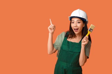 Kadın Asyalı şok ressam boya fırçasıyla turuncu arka planda bir şeyi işaret ediyor.