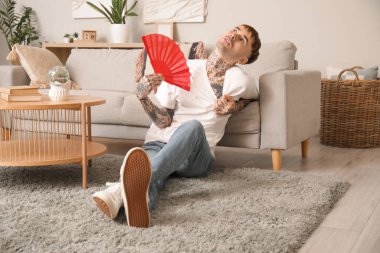 Elinde yelpaze olan dövmeli genç adam evde sıcaktan muzdarip.