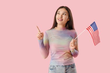 ABD bayrağı taşıyan genç Asyalı kadın pembe arka plandaki bir şeyi işaret ediyor.