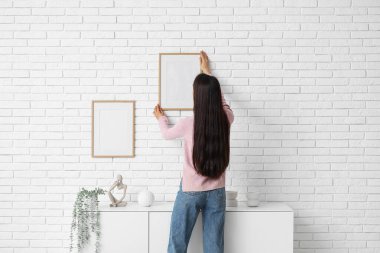 Genç bir kadın odadaki çekmecelerin yanındaki beyaz tuğla duvara resim çerçevesi asıyor.