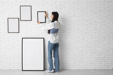 Genç bir kadın şık bir odada beyaz tuğla duvara resim çerçevesi asıyor.