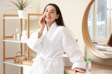 Bornozlu, pamuklu genç ve güzel bir kadın evde makyajını temizliyor.