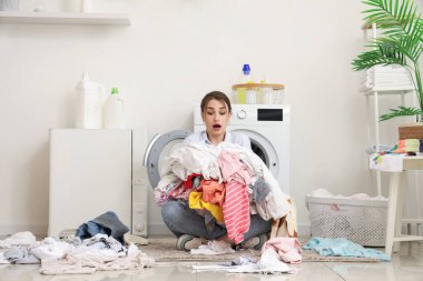 Evde çamaşır makinesinin yanında kirli çamaşır yığınıyla şok olmuş genç bir kadın.