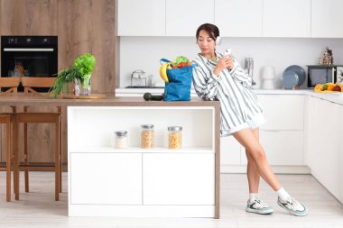 Kulaklıklı genç Asyalı kadın mutfaktaki alışveriş çantasının yanında cep telefonu kullanıyor.