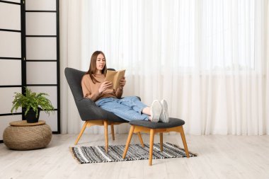 Oturma odasının penceresinin yanındaki rahat koltukta kitap okuyan güzel genç bir kadın.