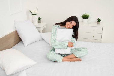 Pijamalı genç bir kadın evde rahat bir yatakta yumuşak ortopedik yastık tutuyor.