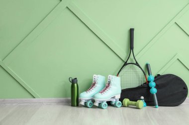 Çantalı tenis raketi, spor su şişesi, dambıl, eski patenler ve yeşil duvarın yanında duran tekerlekli masaj çubuğu.