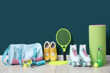 Farklı spor aletleri, patenler ve mavi duvarın yanındaki döşemede spor ayakkabıları.