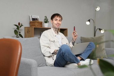 Tek kullanımlık elektronik sigarası ve dizüstü bilgisayarı olan genç adam evdeki kanepede oturuyor.