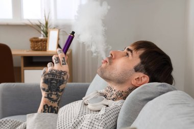 Genç adam tek kullanımlık elektronik sigara içiyor ve evdeki kanepede dinleniyor.