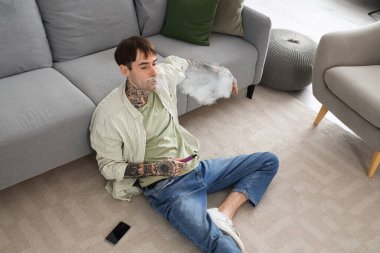 Genç yakışıklı adam tek kullanımlık elektronik sigara içiyor ve evdeki kanepenin yanında oturuyor.