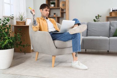 Genç yakışıklı adam tek kullanımlık elektronik sigara içiyor ve evdeki koltukta dizüstü bilgisayar kullanıyor.