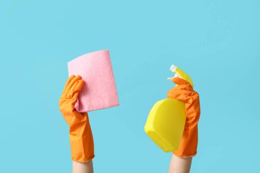 Lastik eldivenli kadın elleri mavi arka planda bez ve bir şişe deterjan tutuyor.