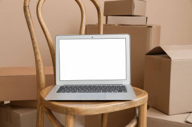 Sandalyedeki boş dizüstü bilgisayar bej duvarın yanındaki kutuları taşıyor, yakın plan.