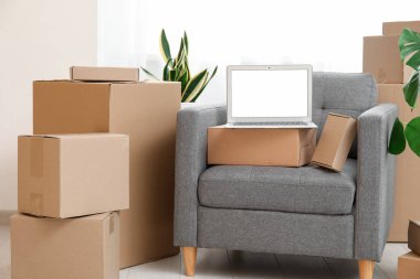 Boş dizüstü bilgisayar, koltukta kutular ve odanın penceresinin yanında bitkiler.