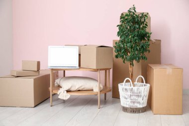 Masada boş dizüstü bilgisayar var. Odada pembe duvarın yanında kutular var.